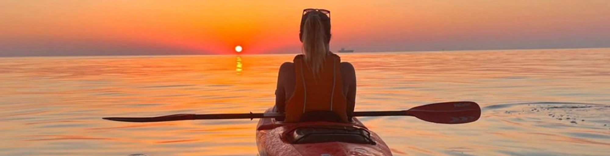 A woman sits in a kayak, facing towards the horizon as the sun sets. She appears contemplative, with a mix of nostalgia and excitement for the new chapter in her life. The vast expanse of water stretches before her, symbolizing the journey ahead filled with decisions and adventures.