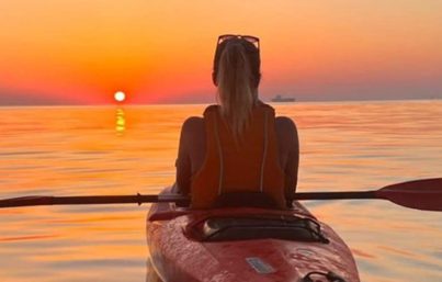 A woman sits in a kayak, facing towards the horizon as the sun sets. She appears contemplative, with a mix of nostalgia and excitement for the new chapter in her life. The vast expanse of water stretches before her, symbolizing the journey ahead filled with decisions and adventures.