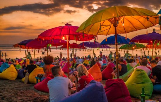 Denpasar, bali, indonesia - 17 june 2018: tourists relaxing and sitting on colorful bean bags, under the umbrellas, and enjoying the sunset at the beach.