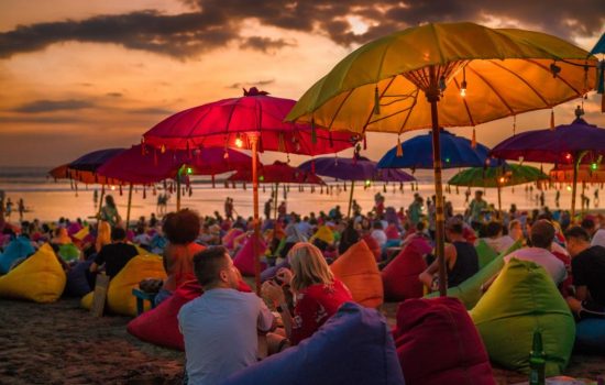Crowded tourists relaxing on the beach at dusk in Bali, Indonesia Denpasar, Bali, Indonesia - 17 June 2018: Tourists relaxing and sitting on colorful bean bags, under the umbrellas, and enjoying the sunset at the beach.